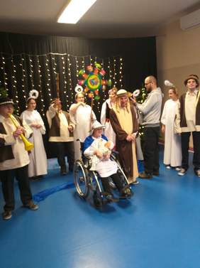 Pokaż zdjęcie: osoby uczestniczą w przedstawieniu dziewczynki są przebrane za anioły a chłopcy w strojach góralskich  śpiewają i tańczą po środku siedzi na wózku chłopak i trzyma niemowle 