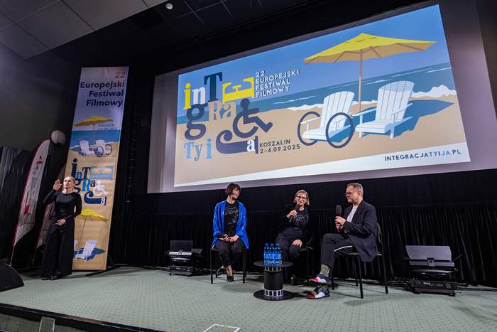 Pokaż zdjęcie: Trzy osoby siedzą na scenie z mikrofonami i rozmawiają podczas spotkania w ramach festiwalu filmowego. Obok nich na ekranie wyświetla się grafika wydarzenia przedstawiająca plażę z leżakami i parasolem oraz logo festiwalu „Integracja Ty i Ja”. Po lewej stronie stoi tłumaczka języka migowego.