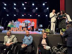 Kadrowanie pokazujące dwa plany wydarzenia. Na podwyższonej scenie teatralnej, po prawej stronie, stoi dwoje prowadzących z mikrofonami i dokumentami (mężczyzna w jasnym garniturze oraz kobieta w ciemnej sukience). W głębi sceny widać grupę osób przy dekoracji z atrap ceglanych murków. Poniżej sceny, na pierwszym planie, siedzi troje uczestników – dwie osoby na wózkach inwalidzkich oraz jedna na krześle, trzymające małe instrumenty perkusyjne (tamburyna). Nad sceną wisi profesjonalne oświetlenie estradowe.