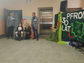 Na tle ściany, oraz otwartych drzwi oraz zamknietych dzwi windy widzimy dwóch młodych mężczyzn między którymi na wózku inwalidzkim siedzi dziewczynka, obok niej przykuca kobieta, z prawej strony na ciemno zielonej tablicy napis 30 lat pfron