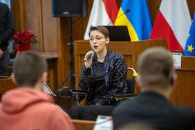 Na środku sali siedzi na wózku inwalidzkim kobieta, trzyma w dłoni mikrofon i śpiewa