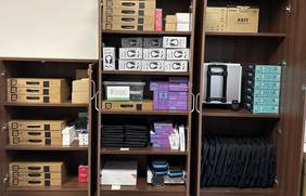 Pokaż zdjęcie: W otwartej, brązowej szafie na półkach stoi sprzęt elektroniczny popakowany w oryginalne opakowania.