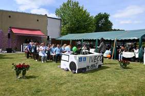 Pokaż zdjęcie: Kilkanaście osób siedzi na krzesłach ustawionych w kilku rzędach, na trawniku. Z przodu konstrukcja z kartonów imitująca maskę autobusu. Na trawniku jest też pięć wazonów z czerwonymi różami. Po prawej stronie długi namiot, pod którym siedzą ludzie.