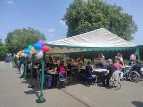 Pokaż zdjęcie: Grupa kilkudziesięciu osób siedzi przy stolikach ustawionych pod dużym namiotem i spożywa posiłek. Namiot udekorowany jest kolorowymi balonami. W tle brama wjazdowa, samochód osobowy, wysokie drzewa liściaste oraz niemal bezchmurne niebo. 