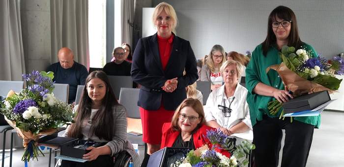 Pokaż zdjęcie: Z przodu sali znajdują się dwie kobiety poruszające się na wózku wraz ze statuetką, teczką oraz bukietem kwiatów, natomiast w drugim rzędzie stoją także dwie kobiety, z czego jedna z nich trzyma te same nagrody. W tle mężczyzna i grupa kilku kobiet.