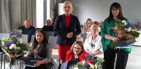 Pokaż zdjęcie: Z przodu sali znajdują się dwie kobiety poruszające się na wózku wraz ze statuetką, teczką oraz bukietem kwiatów, natomiast w drugim rzędzie stoją także dwie kobiety, z czego jedna z nich trzyma te same nagrody. W tle mężczyzna i grupa kilku kobiet.