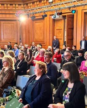 Pokaż zdjęcie: Grupa około trzydziestu osób znajduje się w sali konferencyjnej, której ściany wyłożone są drewnianą boazerią. Z tyłu sali znajdują się reflektory oraz rzutnik.