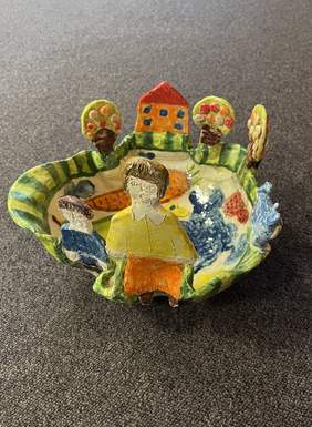 Pokaż zdjęcie: Kolorowa misa z ceramiki, na brzegach wystające dwie postacie, piesek, drzewa owocowe i domek.  Na środku namalowana marchewka, jabłuszko, gruszka, motyl, pszczoła i ryba. 