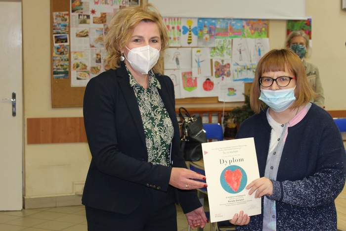 Pokaż zdjęcie: Dwie kobiety w maseczkach trzymające dyplom, na którym widnieje czerwone serce na niebieskim kółku. Za kobietami  tablica ogłoszeń z wywieszonymi kolorowymi pracami.