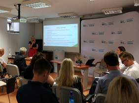 Pokaż zdjęcie: Kobieta stoi z mikrofonem i omawia zmiany w prowadzone do systemu po środku zamieszczono duży ekran na którym zamieszczono slajdy o zmianach w programie Systemu Obsługi Wspracia. Na dużej sali siedzą osoby i słuchają 