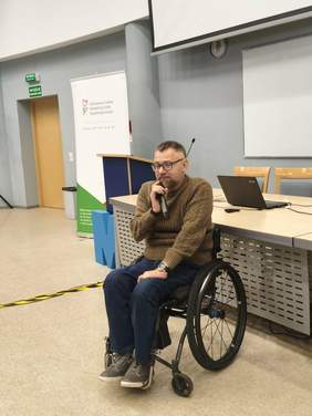 Mężczyzna w brązowym swetrze i czarnych spodniach, siedzi na wózku, w ręce trzyma mikrofon