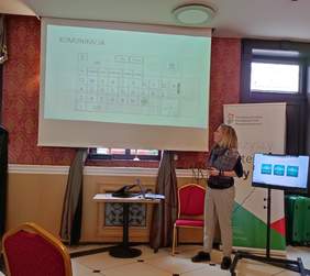 Pokaż zdjęcie: Kobieta z chustą na ramionach stoi przy ekranie na którym wyświetlona jest prezentacja. 