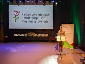 Pokaż zdjęcie: Przy mównicy, na tle logo PFRON, stoi mężczyzna wygłaszający wystąpienie