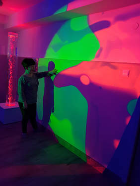 Pokaż zdjęcie: W ciemnym pokoju przy ścianie stoi kobieta oparta ramienbiem o ścianę. Ściana jest w kolorze różowo fioletowym z zieloną plamą na środku. Na ścianie odbija się cien kobiety. Z tyłu w rogu widać podświetloną tubę. anatowym 