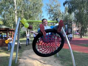 Pokaż zdjęcie: Chłopiec huśta się na huśtawce wahadłowej "bocianie gniazdo". Jest uśmiechnięty. W tle widać inne dzieci bawiące się na placu zabaw. 