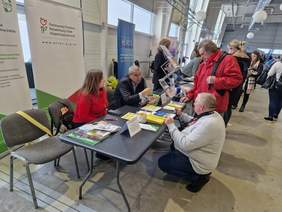 Pokaż zdjęcie: Zdjęcie Targów, widać rząd stolików przy których siedzą wystawcy, naprzeciwko stoją zainteresowani. W tle widać rollupy reklamowe poszczególnych wystawców.