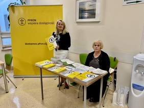 Pokaż zdjęcie: Dwie kobiety, jedna siedzi za biurkiem, druga stoi obok banery z napisem PFRON wspiera i trzyma informator w ręku.