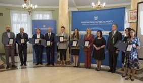 Pokaż zdjęcie: W sali kolumnowej zdjęcie laureatów stojących w półkolu, w rękach prezentują otrzymane dyplomy. W tle banery Państwowej Inspekcji Pracy 