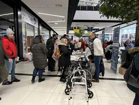 Pokaż zdjęcie: Na pierwszym planie chodzik i wózek inwalidzki, za nimi w szeregu burka przy których znajdują się wystawcy. Przed biurkami dużo ludzi rozmawia lub spogląda na materiały na biurkach.