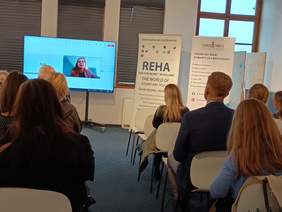 Pokaż zdjęcie: Sala konferencyjna, na białych krzesłach siedzą ludzie którzy patrzą się w wielki telewizor stojący przed nimi, na ekranie pokazywana jest transmisja na żywo kobiety w okularach. Po prawej stronie Reha for The Blind i Fundacji Szansa.