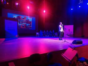 Pokaż zdjęcie: na zdjęciu pięknie oświetlona sala koncertowa w kolrze fioletowo niebieskim, na niej stoi chłopak i śpiewa do widowni ubrany jest w białą koszulę, ma kaszkiet na głowie