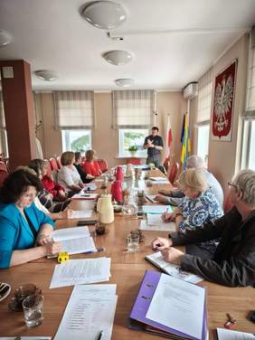 Pokaż zdjęcie: Dziesięć osób siedzi przy dużym stole, na którym znajdują się liczne segregatory, kartki, notesy. Przed nimi stoi mężczyzna w ciemnej koszuli. Na ścianie po prawej stronie duże Godło Polski.