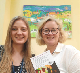 Pokaż zdjęcie: Dwie kobiety uśmiechają się do zdjęcia. Jedna z pań trzyma w dłoni jakąś ulotkę reklamową. W tle widać kolorowy obrazek. 