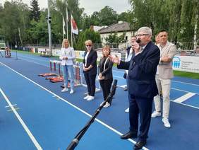 Pokaż zdjęcie: Na boisku sportowym stoją w rzędzie różne osoby. Na pierwszym planie stoi ubrany w garnitur mężczyzna, który przemawia do mikrofonu i wyciąga rękę przed siebie. 