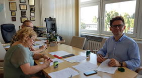 3 osoby przy stole podpisują umowę, od lewej kobieta w zielonej sukience pochyla się i pisze, za nią widać fragment sylwetki mężczyzny, z prawej strony mężczyzna w niebieskiej koszuli patrzy w obiektyw, usmiecha się