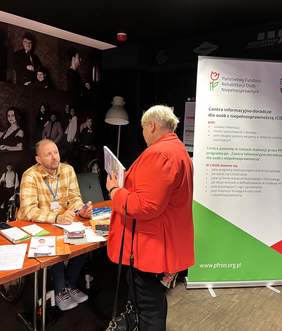 Stoisko PFRON - za stołem siedzi przedstawiciel PFRON - Paweł Dzik, a przed nim stoi kobieta. Po prawej stronie widoczny baner PFRON-u