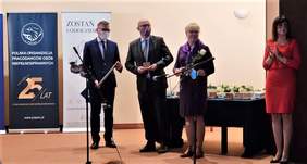 Pokaż zdjęcie: W Auli na scenie stoją dwaj mężczyźni i dwie kobiety, wśród nich dyrektor Marta Mordarska. Czekają na Laureatów, którym wręczać będą nagrody. Obok nich stolik z przygotowanymi nagrodami