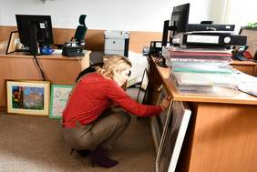 Pokój, o biurka oparte prace. Pani Bogna Jarzemska-Misztalska, kucając przygląda się jednej z ocenianych prac. 