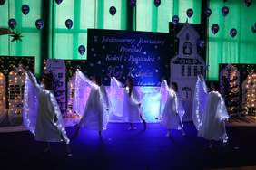 Pokaż zdjęcie: Na scenie występuje grupa dziewcząt ubranych w białe, anielskie stroje z podświetlanymi skrzydłami. Tancerki wykonują układ taneczny w świątecznej scenerii z choinkami, świetlnymi dekoracjami i banerem „X Jubileuszowy Powiatowy Przegląd Kolęd i Pastorałek Hej, Kolęda” w tle.