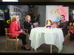 Pokaż zdjęcie: Przy okrągłym stole z białym obrusem siedzą trzy kobiety. Jedna z nich trzyma w ręku mikrofon i przeprowadza wywiad. Za paniami stoi mapa Ukrainy