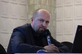 Pokaż zdjęcie: Zdjęcie łysiejącego mężczyzny z lekka brodą podczas prezentacji. Mężczyzna trzyma w ręku mikrofon i spogląda na laptopa