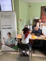 Kobieta stoi przy stoliku naprzeciw dwóch siedzących mężczyzn. Na stole leżą dokumenty i materiały informacyjne, a w tle widoczny jest kolejny mężczyzna stojący obok stanowiska.