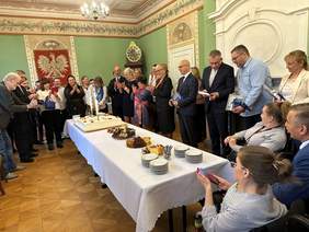 Pokaż zdjęcie: W eleganckiej sali muzealnej dookoła długiego stołu stoją ludzie. Na środku stołu znajduje się duży kwadratowy tort z dwiema palącymi się racami. Wszyscy się uśmiechają. W tle godło Polski.