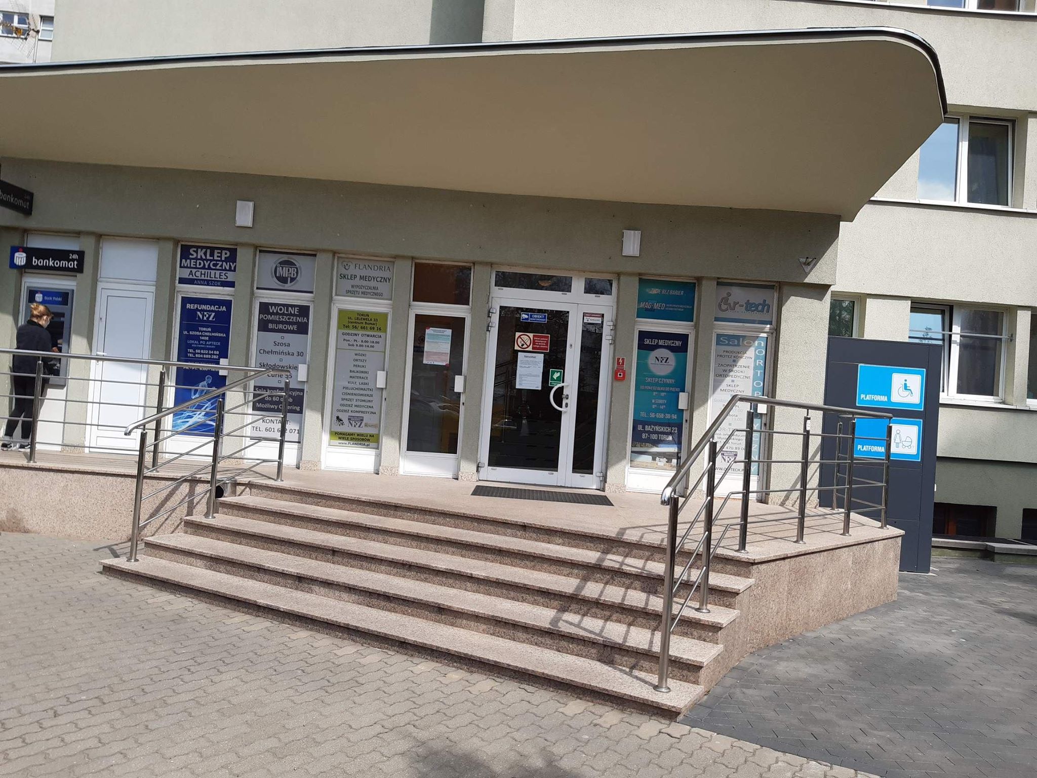 Wejście główne do budynku Wejście do budynku zadaszone asymetrycznym daszkiem, rozciągającym się nad podestem o tym samym kształcie. Na podest którego prowadzą szerokie schody o czterech stopniach, z obydwu stron ograniczone stalowymi poręczami. Na podeście z lewej strony bankomat, przed którym stoi kobieta. Patrząc w praw widzimy szereg reklam instytucji znajdujących się w budynku. W centrum podestu białe drzwi wejściowe do budynku. Po lewej stronie od podestu widać okna budynku i platformę dla wózków inwalidzkich., oznaczoną dużymi napisami i piktogramami na niebieskim tle.