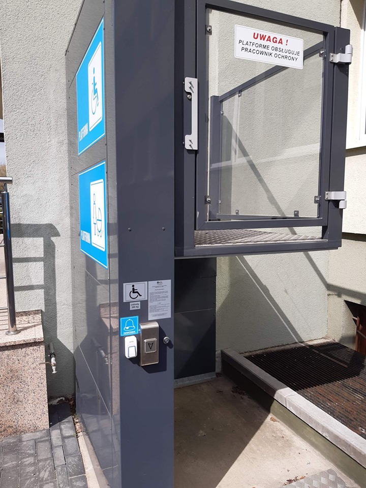 Platforma dla wózków Platforma dla osób na wózkach inwalidzkich w ciemnoszarym kolorze z podjazdem zabezpieczonym szklanymi drzwiami. Na obudowie są tabliczki z piktogramami osób z wózkiem dziecięcym oraz osób z wózkiem dla osób niepełnosprawnych i napisem platforma. Na frontowej ściance oznaczenie z piktogramem osoby niepełnosprawnej oraz napisem „udźwig 385 kg”. Poniżej umieszczono przyciski, jeden z nich jest oznaczony piktogramem dzwonka i służy wzywaniu pomocy, a drugi służy do sterowania platformą. Platforma zatrzymana w pozycji górnej, a na jej drzwiczkach napis „Uwaga, platformę obsługuje pracownik ochrony”.