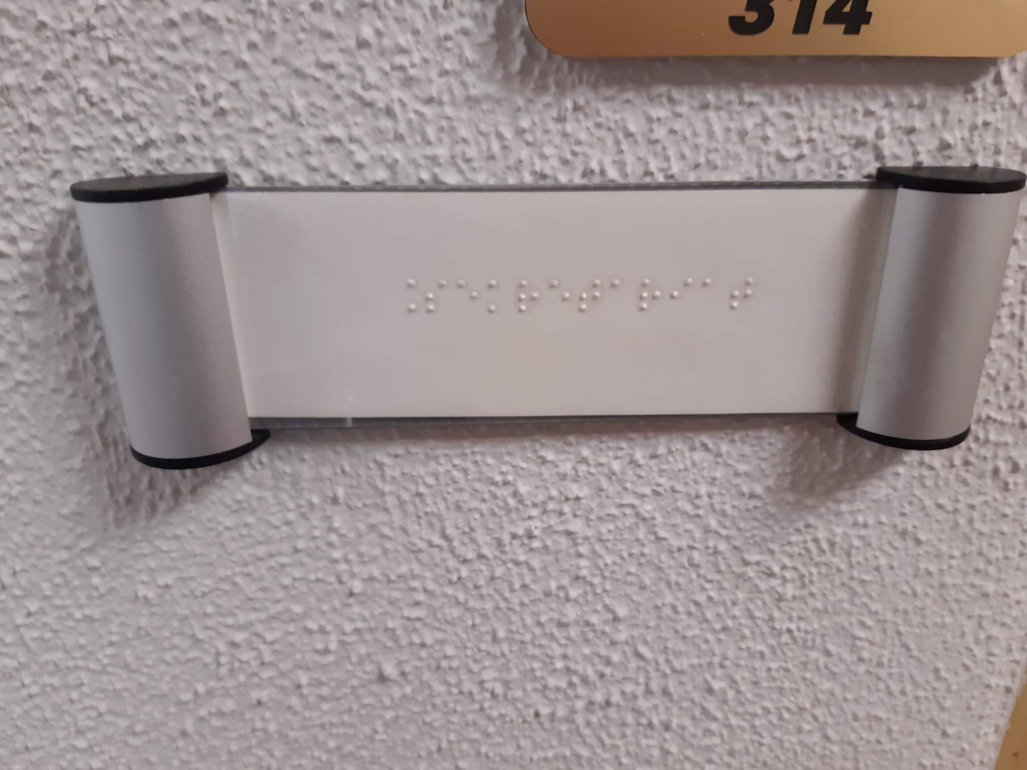 Na jasnej ścianie jasnoszara tabliczka z napisem w języku Braille’a. 