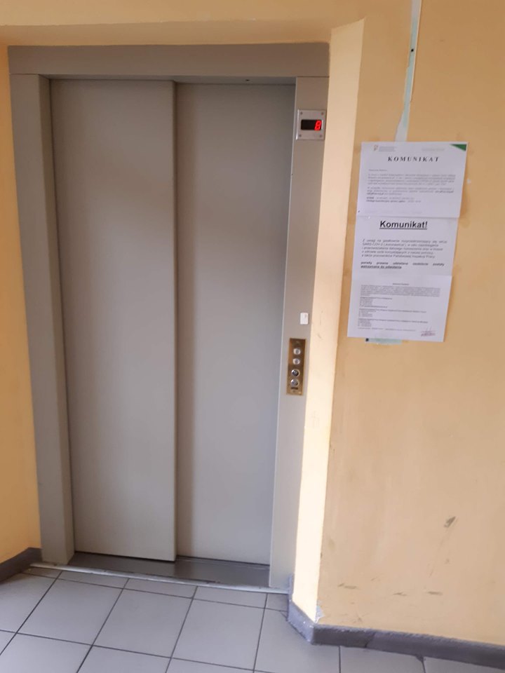 Wejście do windy Pomarańczowa ściana, pośrodku wnęka na drzwi windy - jasnoszare, dwudzielne. Po prawej stronie widzimy panel przyzywania windy. Po prawej stronie od wnęki z drzwiami windy, na ścianie, na jasnych kartkach wydrukowane komunikaty objaśniające wzywanie windy oraz zachowanie w sytuacji pandemicznej