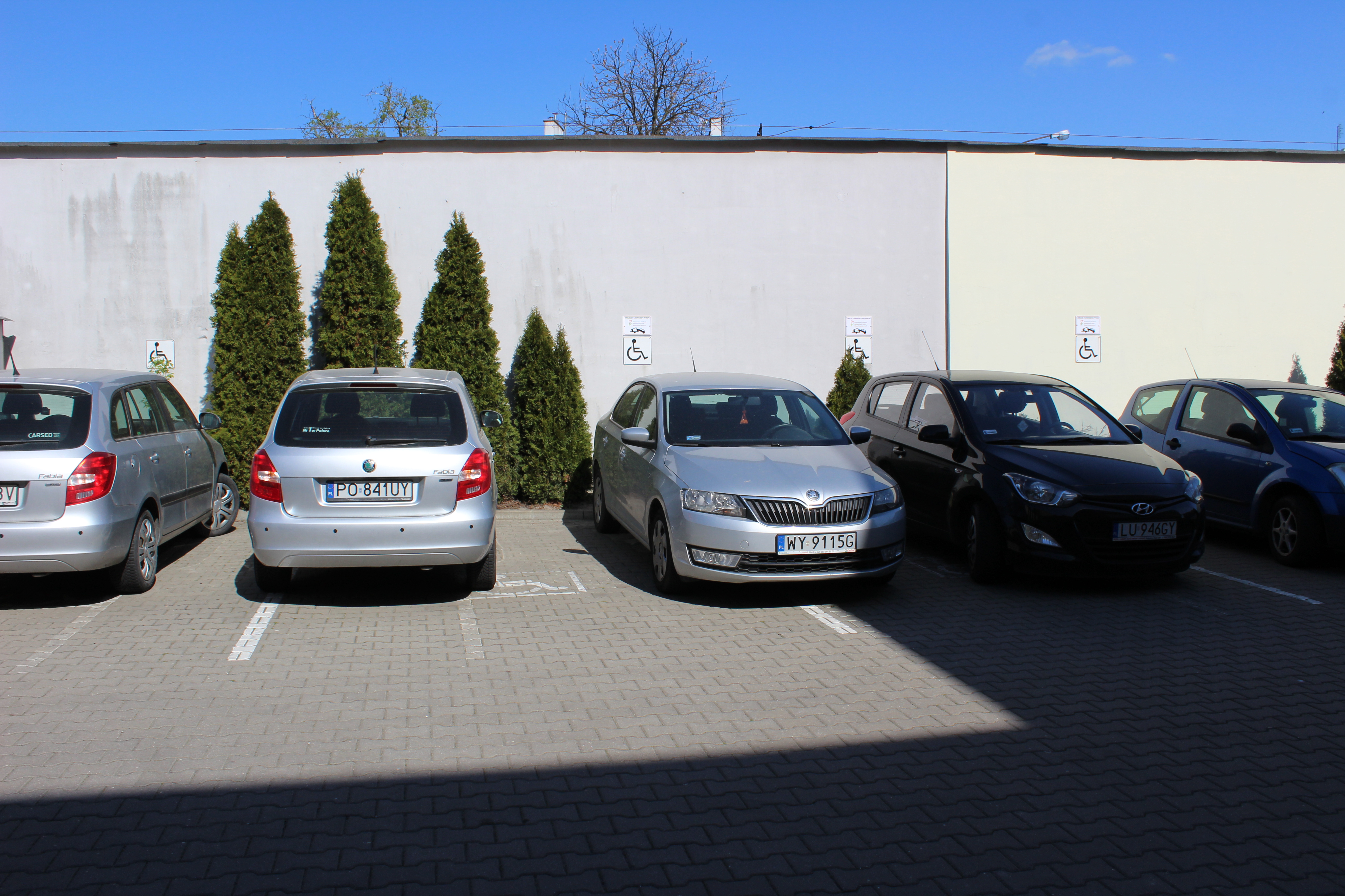 Miejsca parkingowe z zaparkowanymi samochodami na tle jasnożółtej ściany przesłoniętej krzewami