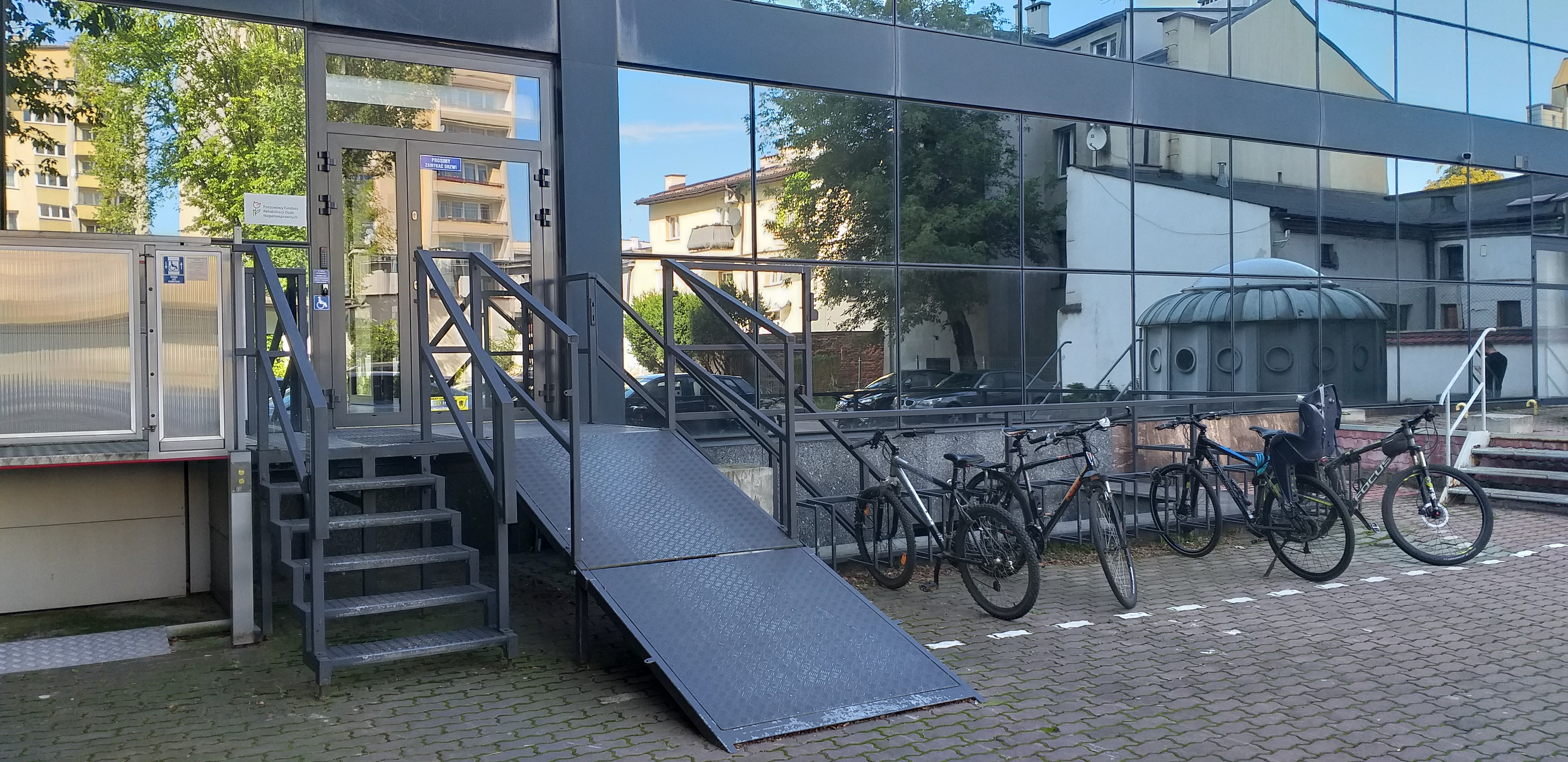 Wejście od parkingu Widok na wejście, do którego prowadzą ażurowe metalowe schody obok których mamy rozkładaną pochylnie dla wózków. Po prawej stronie od pochylni parking dla rowerów. Na wysokości podestu, do którego prowadzą schody i pochylnia, drzwi wejściowe. Na prawo od drzwi szklana ściana, w której odbijają się inne budynki i zaparkowane samochody. Po lewej stronie od schodów widać platformę windy dla wózków