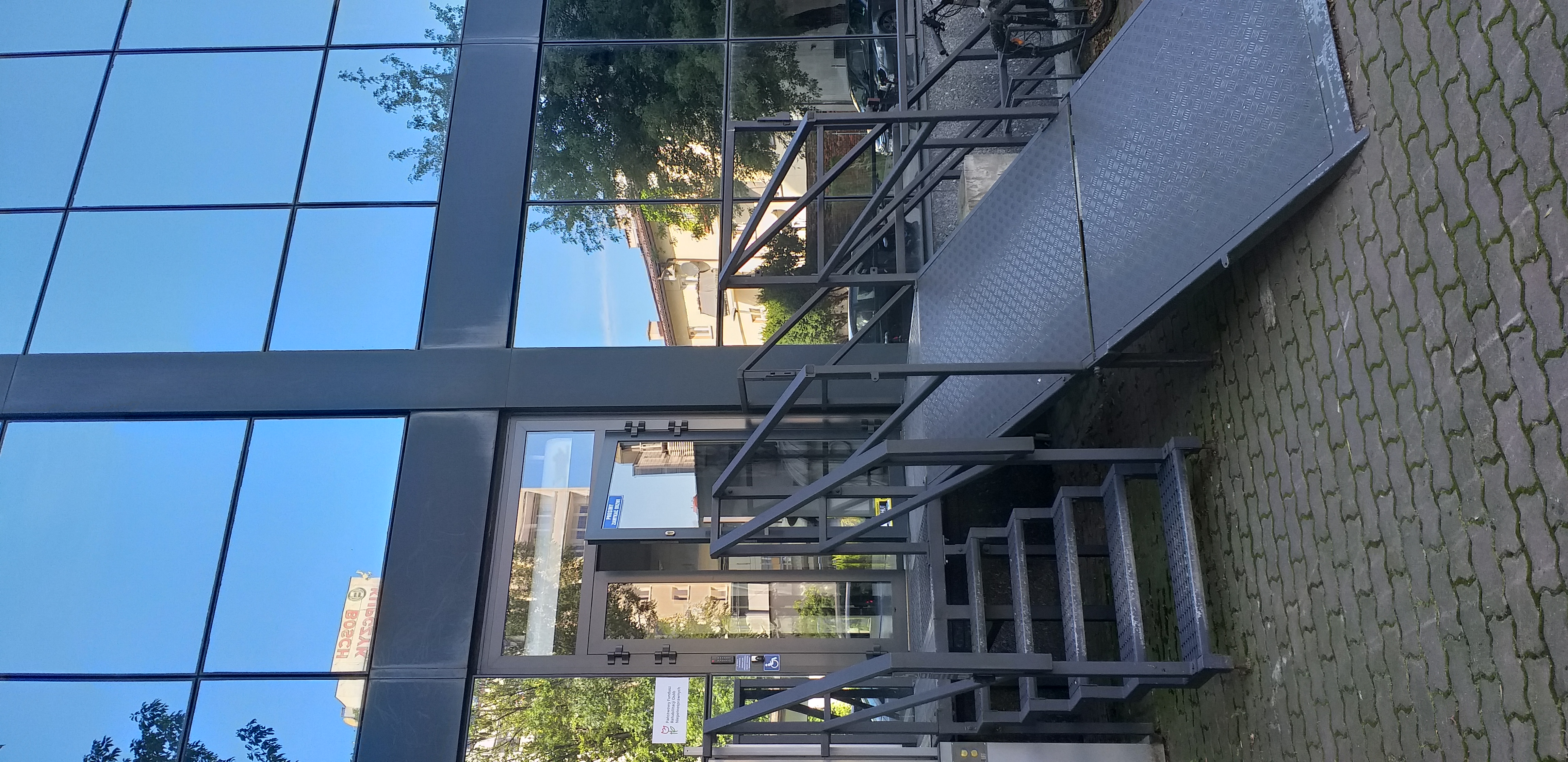 Wejście od parkingu - pochylnia Metalowe, ażurowe schody oraz rozkładana pochylnia prowadzące do drzwi wejściowych do budynku. Budynek cały oszklony, a w jego szklanej ścianie odbijają się drzewa oraz budynki mieszkalne