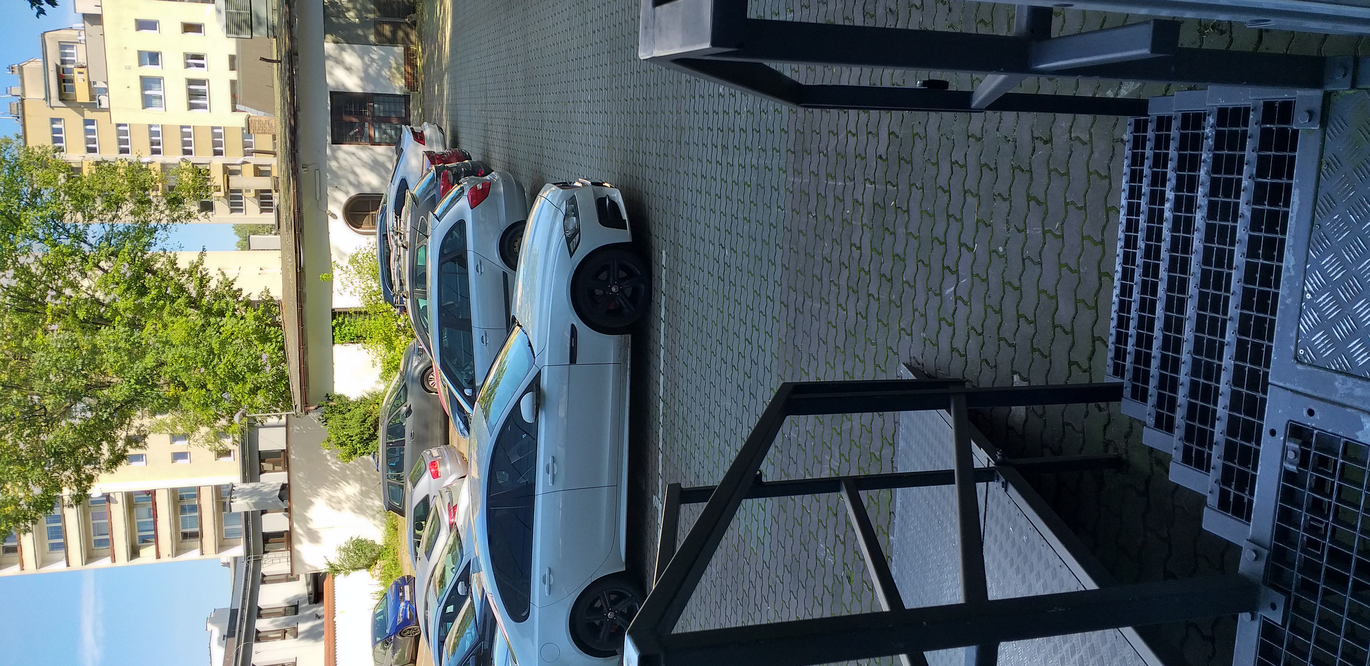 Widok parkingu Widok z drzwi na parking. na pierwszym planie widać schody ażurowe; po ich lewej stronie rozkładana pochylnia. Parking jest otoczony parterowym budynkiem oraz murem. Na parkingu zaparkowane samochody. Za ogrodzeniem parkingu widać wysokie drzewa, a w tle wieżowce mieszkalne