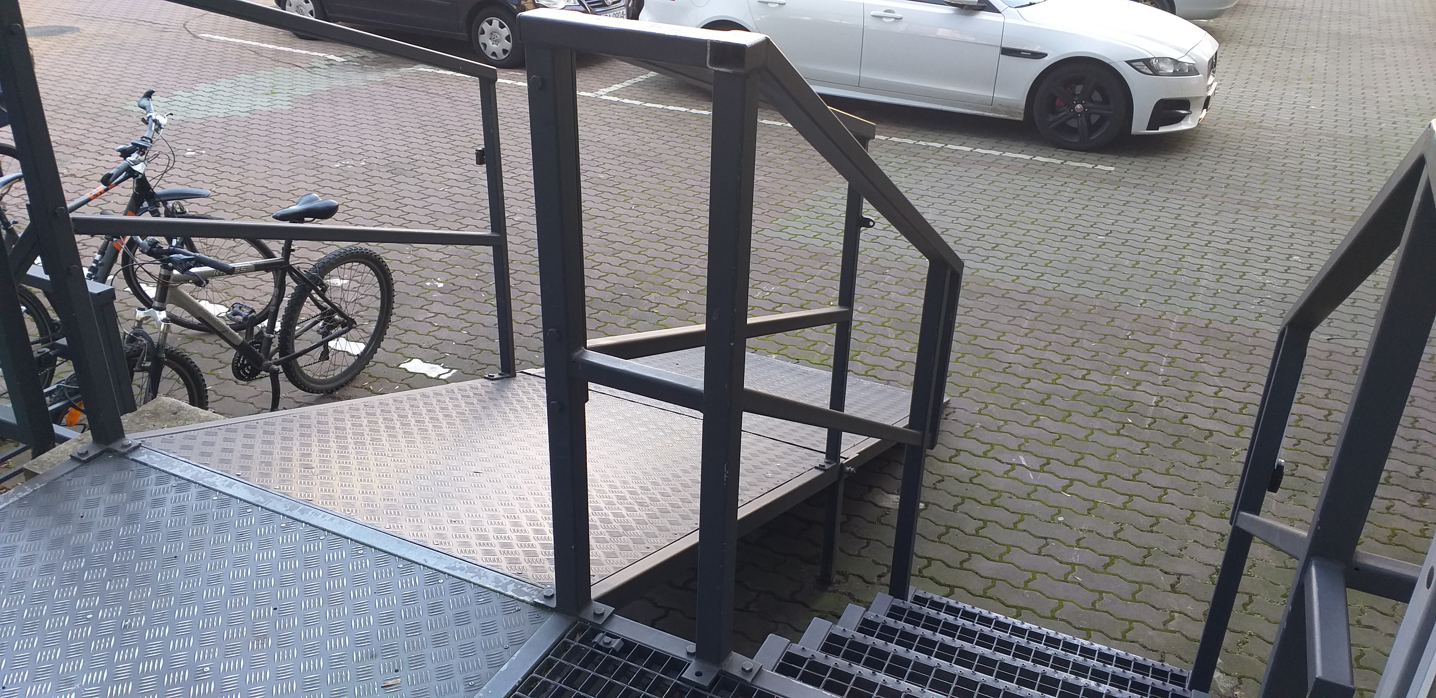 Widok na parking - schody Widok z drzwi wejściowych na rozkładaną pochylnie dla wózków. Po prawej stronie widać ażurowe schody, a po lewej zaparkowane rowery. W tle zaparkowane samochody na parkingu za budynkiem