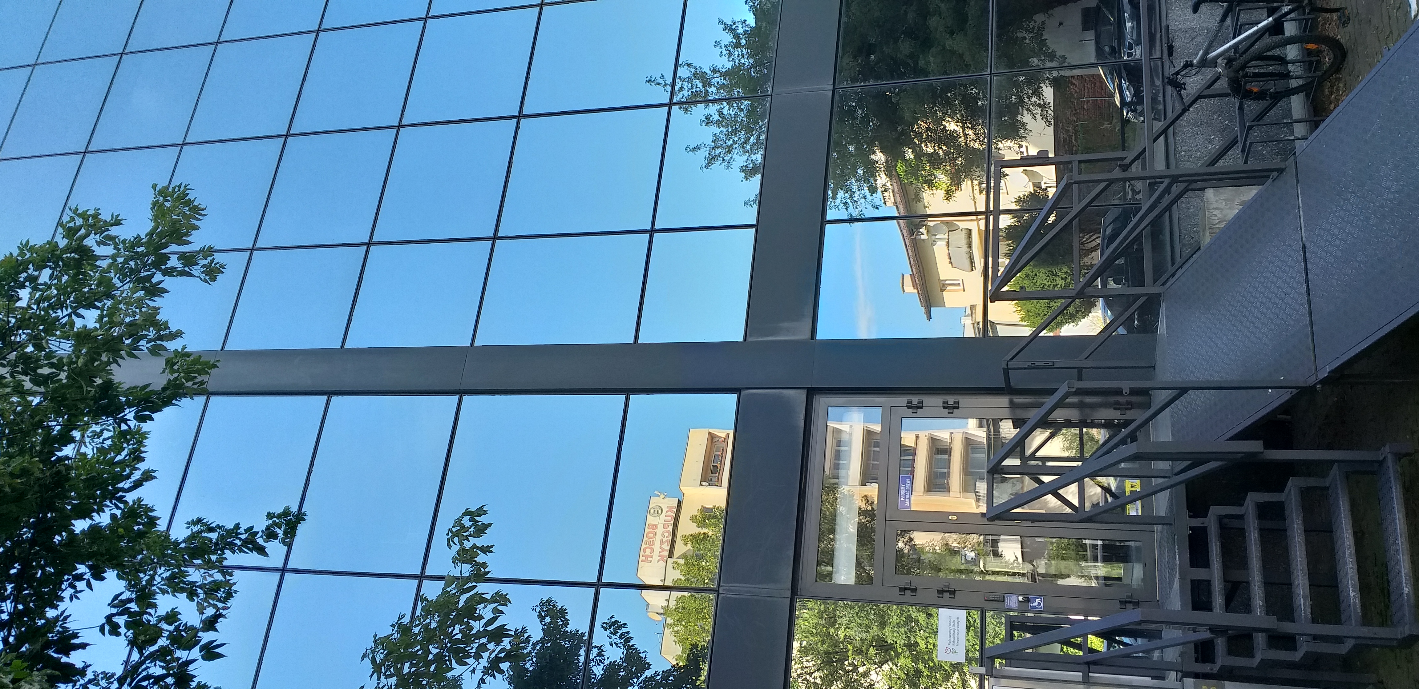 Widok wejścia od parkingu Widok wejścia do budynku. Metalowe schody ażurowej konstrukcji oraz rozkładana pochylnia, wykonana z blachy prowadzą do drzwi wejściowych. Nad drzwiami po ich prawej stronie szklana ściana, w której odbija się okoliczna zabudowa