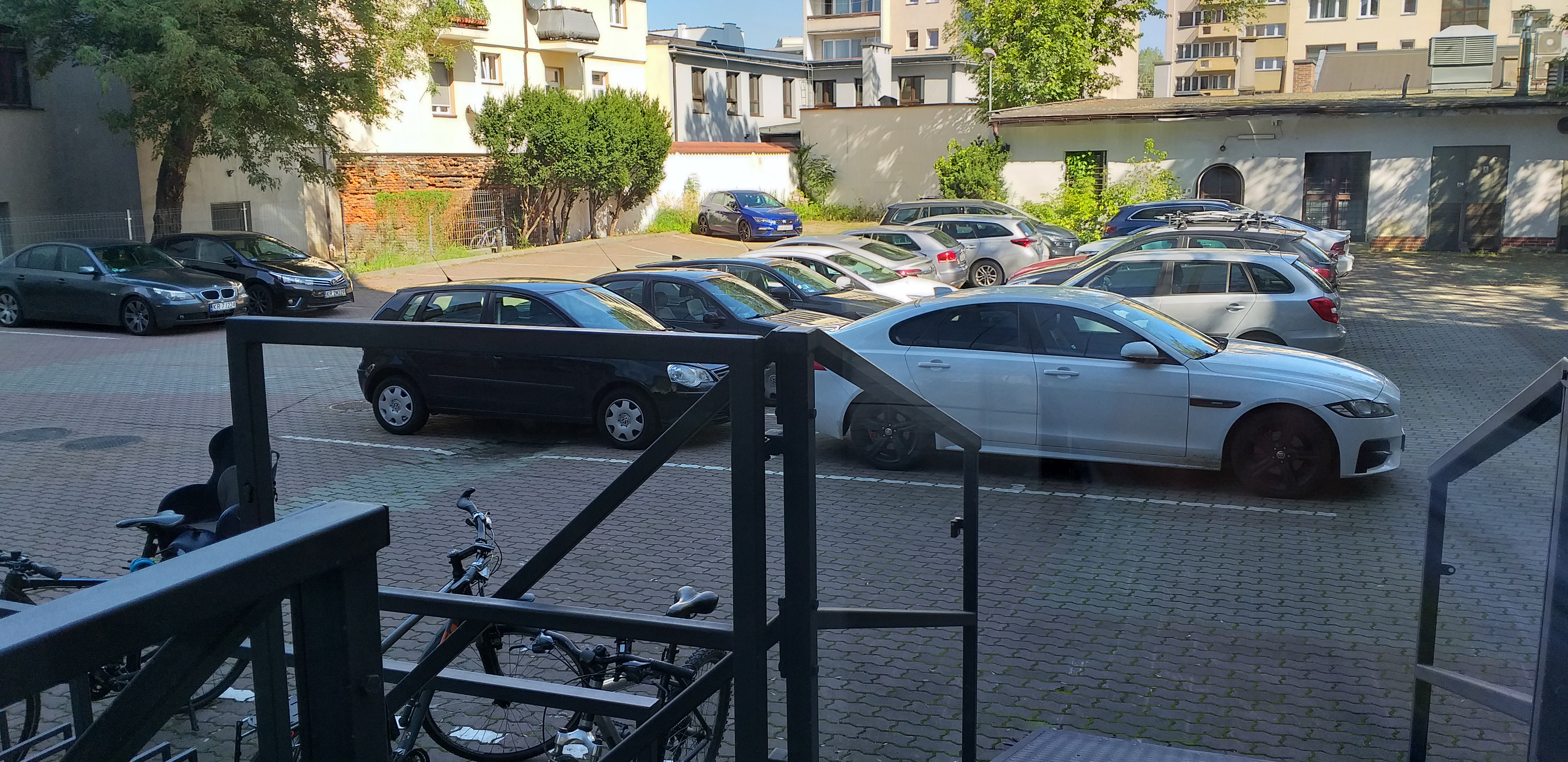 Widok z drzwi wejściowych do budynku na parking. Na pierwszym planie konstrukcja pochylni oraz zaparkowane rowery. Na parkingu zaparkowane samochody, w tle ogrodzenie parkingu oraz budynki parterowe. Za nimi widać rosnące drzewa i budynki mieszkalne 
