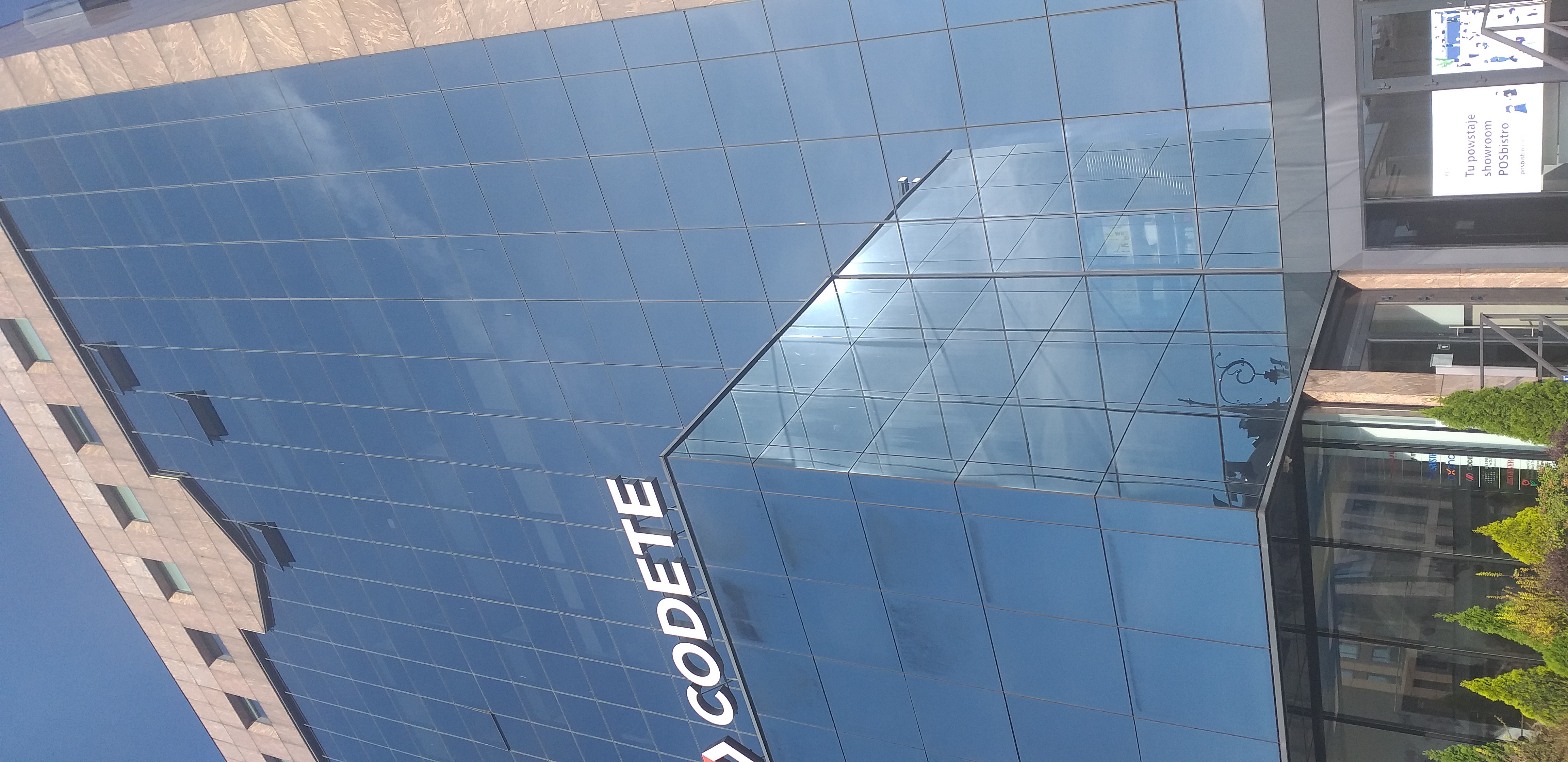 Budynek biurowy ze szklaną ścianą, w której odbija się bezchmurne niebo. Na pierwszym planie niższa część budynku, na której dachu umieszczono napis „Codete”. W prawym, dolnym rogu widać drzwi wejściowe do budynku.