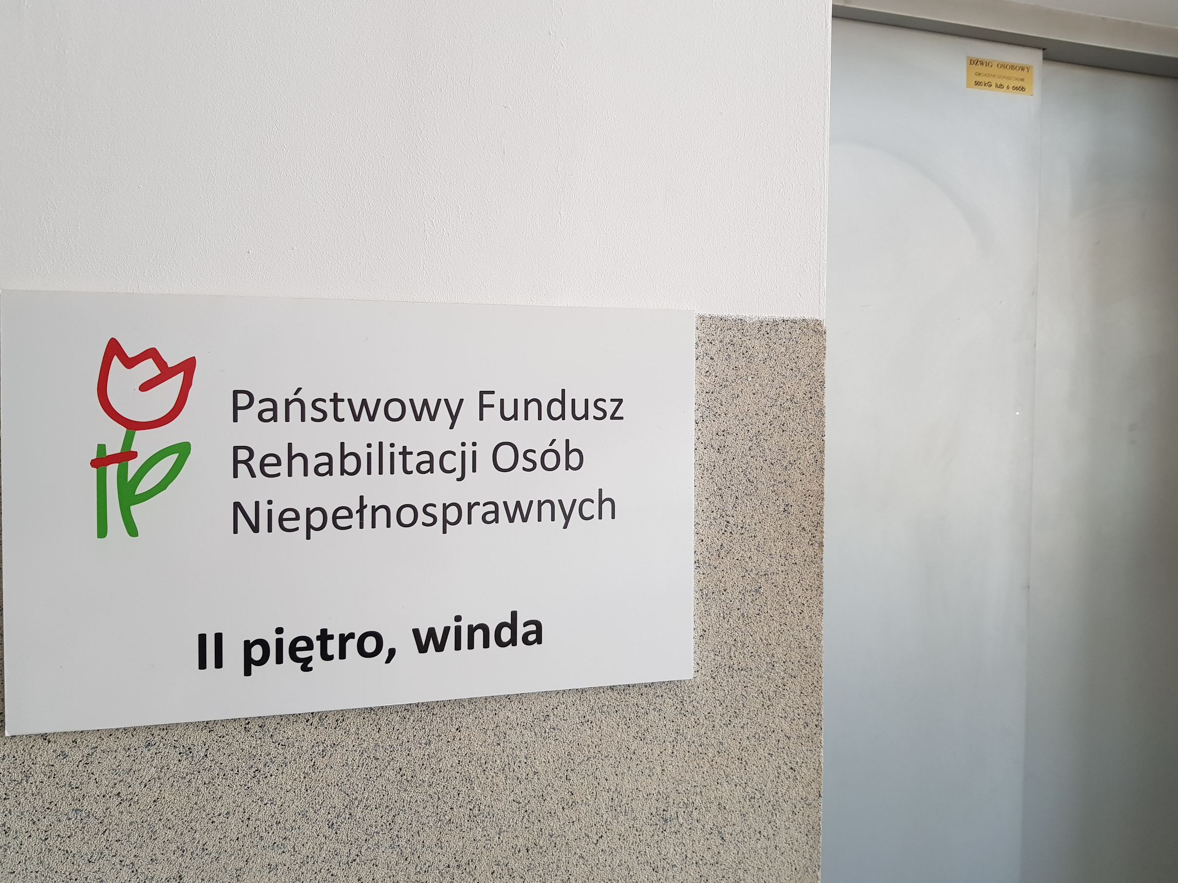 Na białej ścianie nad beżowymi lamperiami wisi tablica informacyjna z logo PFRON i informacją, gdzie mieści się Oddział - drugie piętro, dostępne za pomocą windy. Po prawej stronie od tablicy widoczne drzwi kabiny windy.
